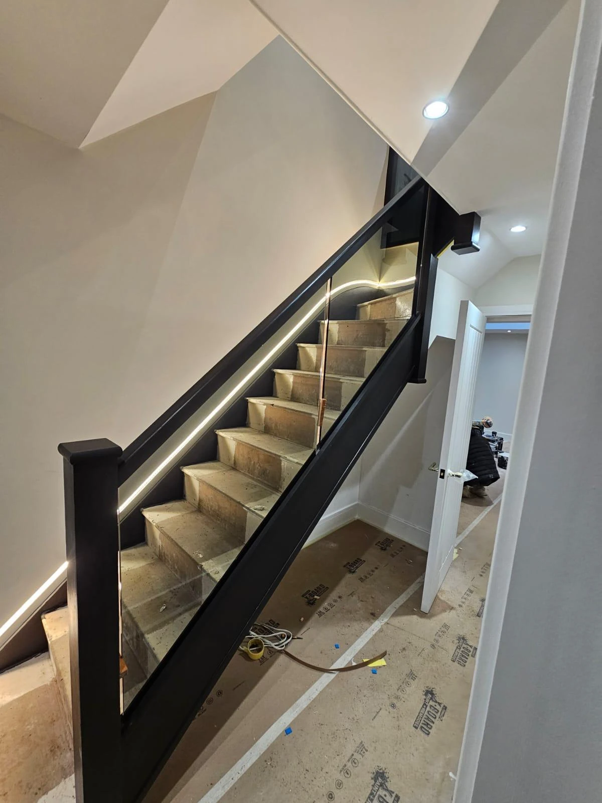 Staircase with modern black railing, illuminated steps, and unfinished flooring. Doors and construction materials visible in the background.